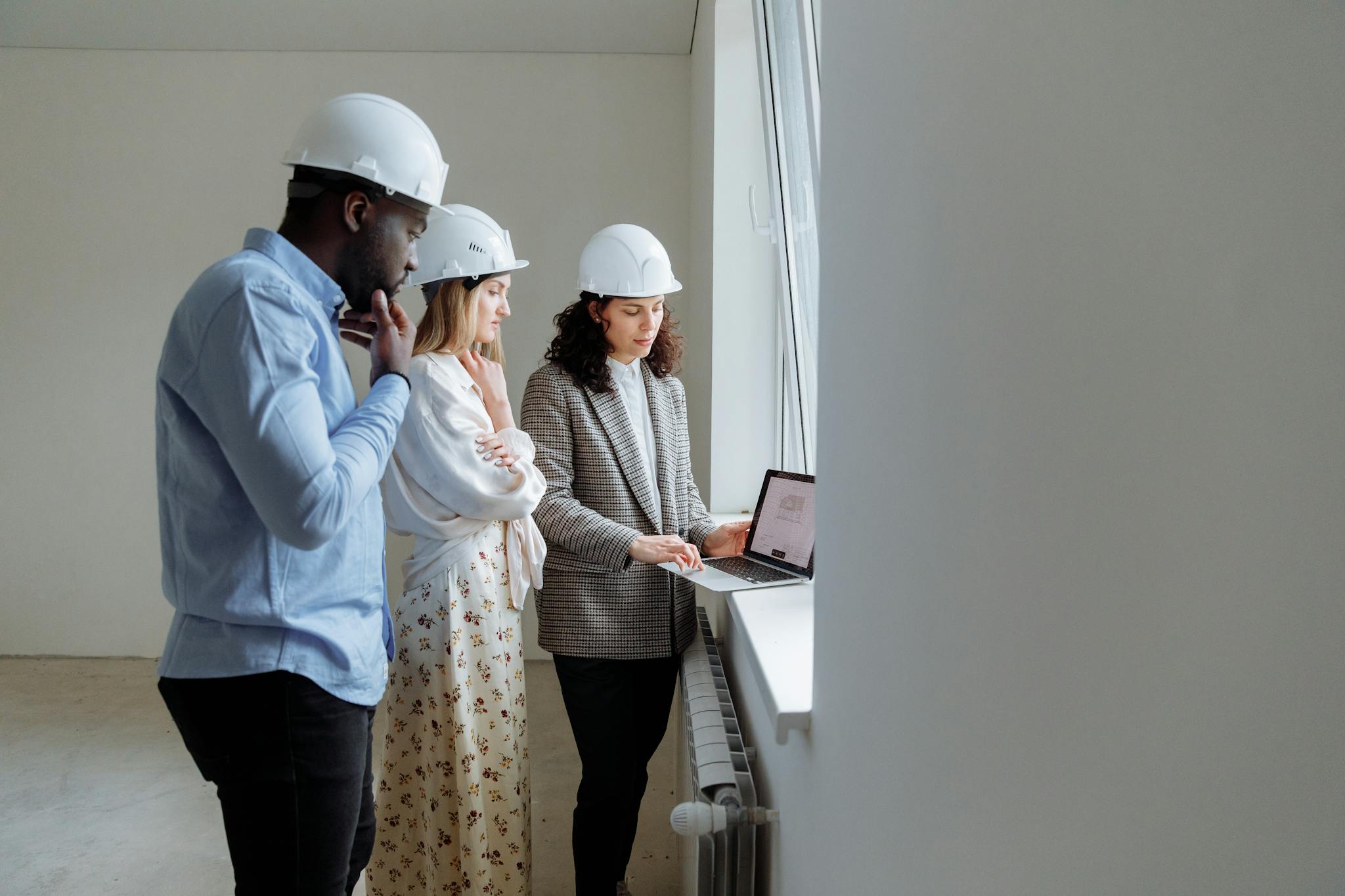 Three engineers wearing helmets review building plans on a laptop in a bright indoor setting.